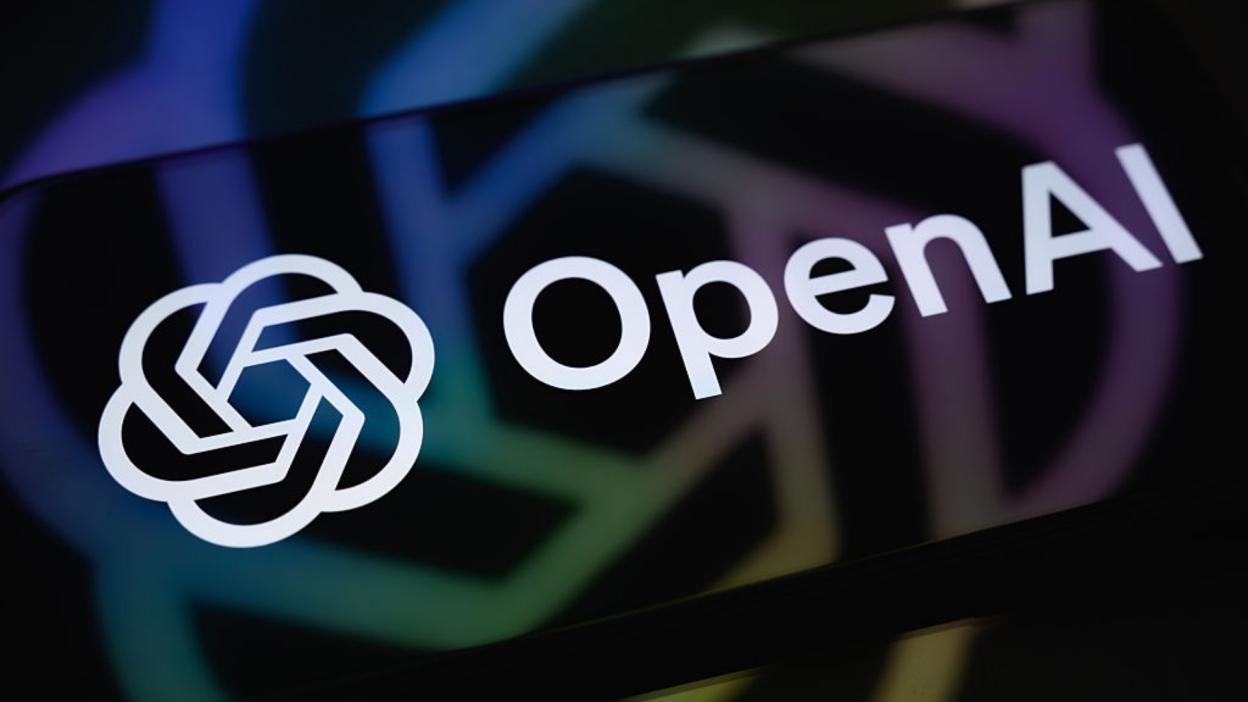 The OpenAI logo is displayed on a smartphone screen resting on a reflective surface with the company